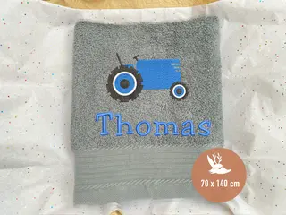 Graues Handtuch mit blauem Traktor-Stickmotiv und personalisierter Bestickung von HeldenFaden. Ideal für Kinder und personalisierte Geschenke.