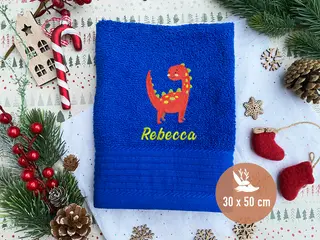 Blaues Handtuch (30x50 cm) mit rotem Dino-Stickmotiv und personalisierter grüner Schrift, umgeben von Weihnachtsdekoration.
