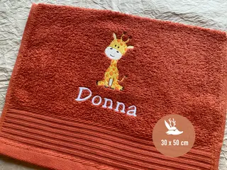 Nahaufnahme eines Handtuchs in der Farbe Terra Spice (30x50 cm) mit Giraffenstickerei und weißem Schriftzug 'Donna'. Ideal für individuelle Geschenke.