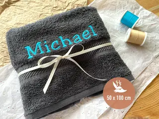 Dunkelgraues Handtuch (50x100 cm) mit dem Namen „Michael“ in türkisfarbener Schrift und weißer Schleife. Elegantes, personalisiertes Geschenk von HeldenFaden.
