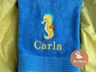 Blaues Handtuch (50x100 cm) mit Seepferdchen-Stickmotiv und dem Namen „Carla“ in gelber Schrift. Hochwertiges, individuell bestickbares Handtuch von HeldenFaden.